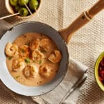Scampis cuits dans une sauce crémeuse servis dans une poêle, accompagnés d’olives et pain à tremper.