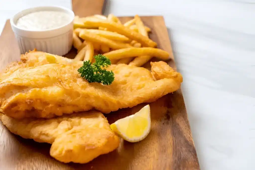 Fish and chips doré servi sur une planche en bois avec des frites, du citron et de la sauce tartare.