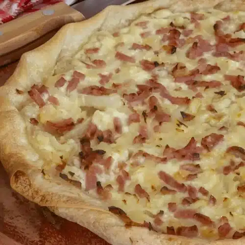 Une délicieuse tarte flambée alsacienne, garnie de crème fraîche, d'oignons et de lardons fumés, présentée sur une planche en bois.