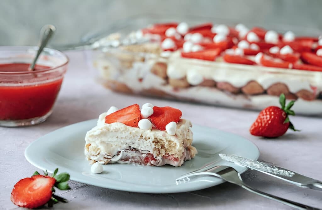 Part de tiramisu aux fraises et mascarpone servie sur une assiette, avec un plat en arrière-plan